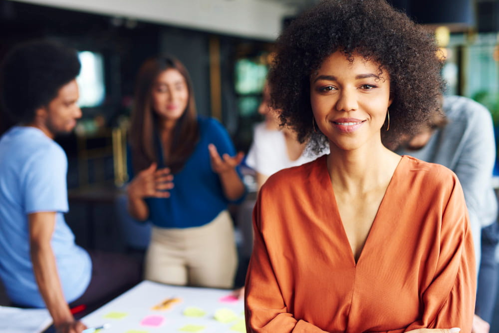 mulher negra sorridente de braços cruzados com um grupo de colegas profissionais em reunião ao fundo