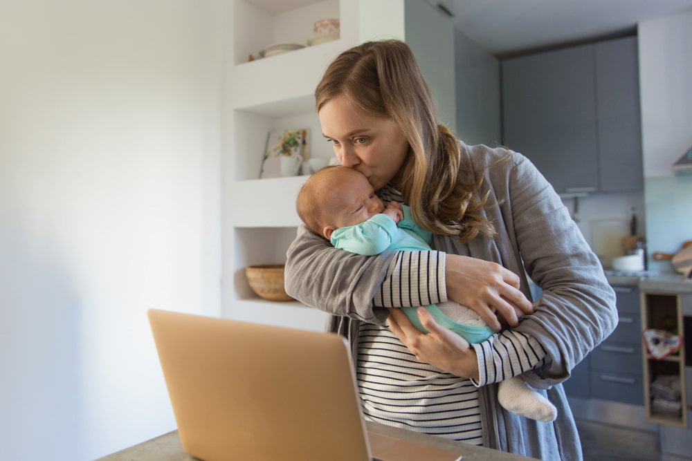 mulher de pé segura um bebê enquanto beija sua testa e olha para a tela de um notebook.